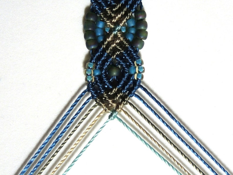 May include: A close-up of a blue, brown, and green macrame bracelet with beads. The bracelet is made with a variety of different colored cords and beads.