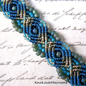 May include: A blue and gray macrame bracelet with turquoise and green beads. The bracelet is made with a knotting technique and features a geometric pattern.