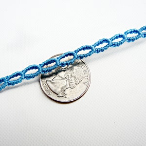 May include: A close-up of a light blue cord bracelet with a knotted design. The bracelet is laid on a white surface next to a US quarter.