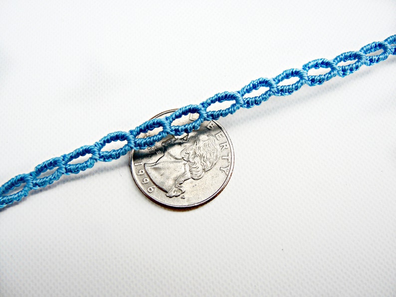 May include: A close-up of a light blue cord bracelet with a knotted design. The bracelet is laid on a white surface next to a US quarter.