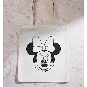 Peut inclure: Un sac fourre-tout en toile blanche avec une illustration en noir et blanc du visage de Minnie Mouse. Minnie Mouse porte un grand n&oelig;ud.