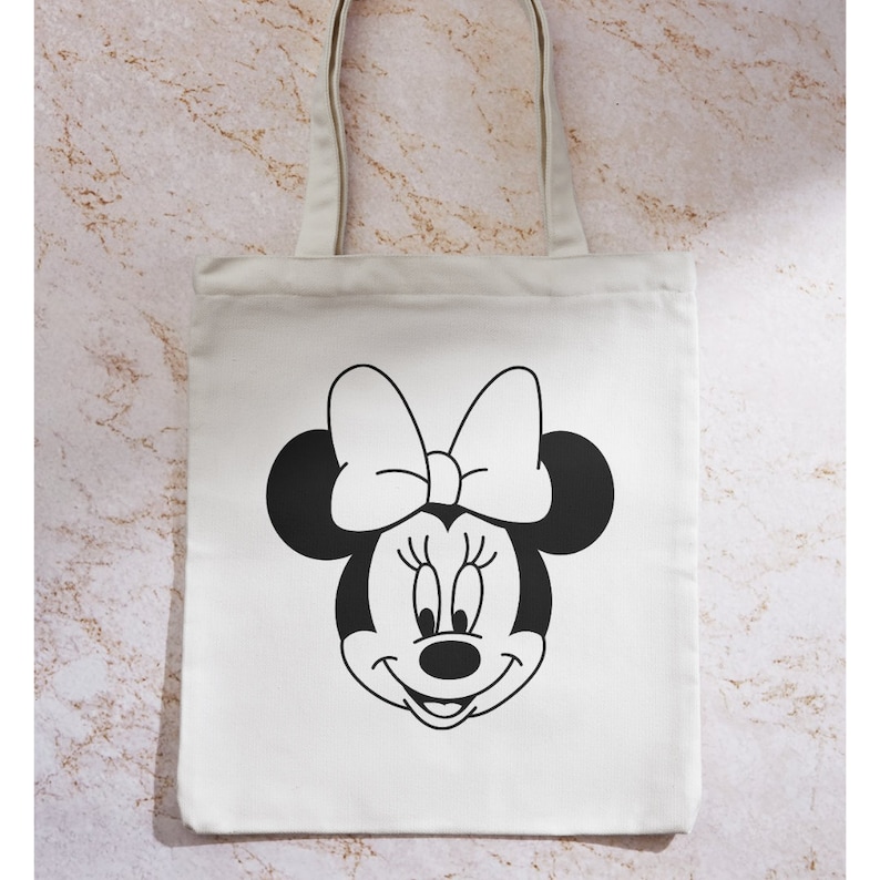 Peut inclure: Un sac fourre-tout en toile blanche avec une illustration en noir et blanc du visage de Minnie Mouse. Minnie Mouse porte un grand n&oelig;ud.