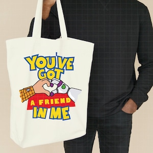 May include: White canvas tote bag with a colorful graphic of Woody and Buzz Lightyear from Toy Story. The graphic says "You've Got A Friend In Me".