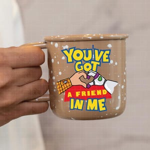 May include: Brown enamel mug with a white speckled design. The mug features a cartoon image of Woody and Buzz Lightyear from Toy Story holding hands in the shape of a heart. The text "You've Got A Friend In Me" is printed on the mug.