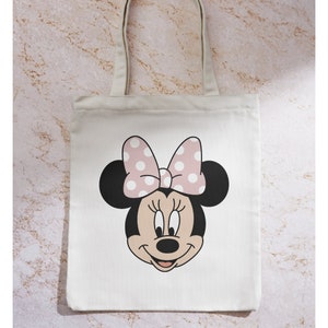 Peut inclure: Un sac fourre-tout en toile blanche avec un n&oelig;ud rose &agrave; pois blancs et une image de dessin anim&eacute; du visage de Minnie Mouse.
