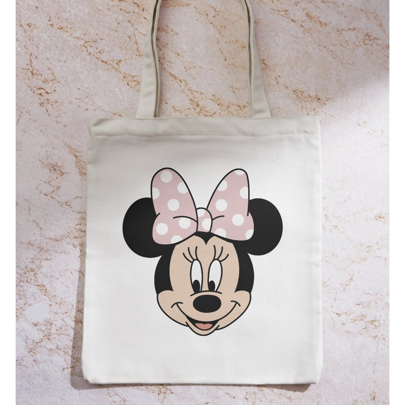 Peut inclure: Un sac fourre-tout en toile blanche avec un n&oelig;ud rose &agrave; pois blancs et une image de dessin anim&eacute; du visage de Minnie Mouse.