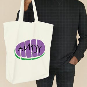 May include: White canvas tote bag with purple and green graphic of a character from the movie Toy Story. The graphic says "ANDY" in black lettering.