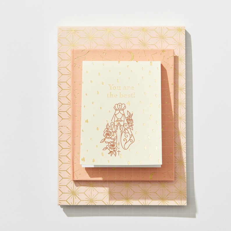 May include: A stack of three notebooks with a gold geometric pattern on the cover. The top notebook has a white cover with a gold illustration of a woman in a veil and the text "You are the best!"