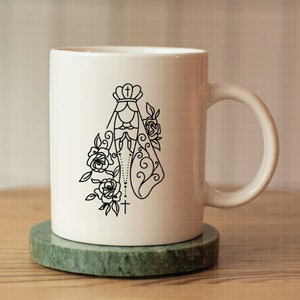 May include: White ceramic mug with a black line drawing of the Virgin Mary with a crown, rosary beads, and flowers. The mug has a handle.