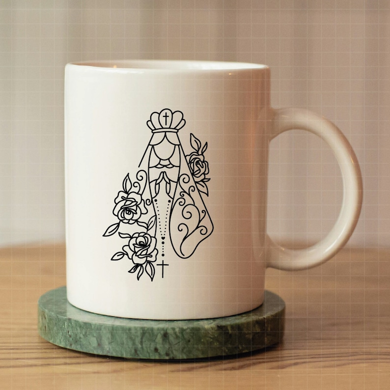 May include: White ceramic mug with a black line drawing of the Virgin Mary with a crown, rosary beads, and flowers. The mug has a handle.