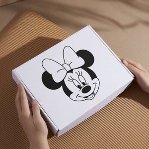 Peut inclure: Une bo&icirc;te en carton blanche avec une illustration en noir et blanc de Minnie Mouse sur le devant. Minnie Mouse porte un n&oelig;ud et a un grand sourire.