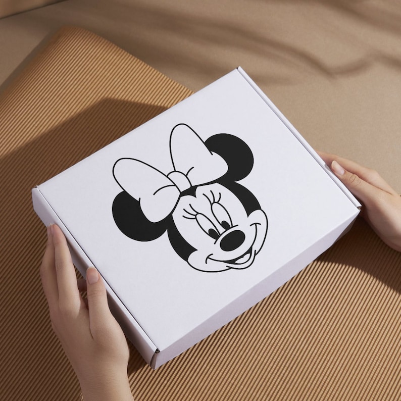 Peut inclure: Une bo&icirc;te en carton blanche avec une illustration en noir et blanc de Minnie Mouse sur le devant. Minnie Mouse porte un n&oelig;ud et a un grand sourire.