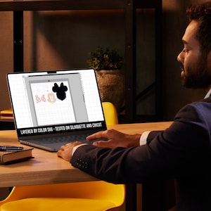 Peut inclure: Une personne utilise un ordinateur portable pour concevoir un fichier SVG en couches. Le design pr&eacute;sente une silhouette noire de la t&ecirc;te de Mickey Mouse avec des accents roses et blancs. Le texte &agrave; l'&eacute;cran indique "Layered by Color SVG - Tested on Silhouette and Cricut".