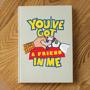 May include: A white notebook with a cartoon image of Woody and Buzz Lightyear from Toy Story. The image shows them holding hands in the shape of a heart. The text "You've Got A Friend In Me" is printed in blue, yellow, and red letters.