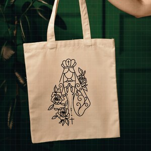 May include: A white canvas tote bag with a black line drawing of the Virgin Mary with a crown and rosary beads. The drawing is surrounded by roses.