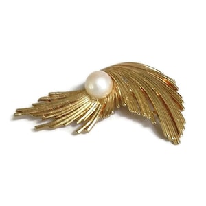 May include: A gold-toned brooch with a single white pearl in the center. The brooch is shaped like a stylized feather with a curved design.