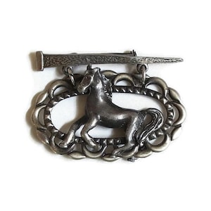 May include: A silver-toned brooch featuring a horse within an oval frame, with a textured nail design above. The horse is in a running pose. The brooch has a pin closure on the back.