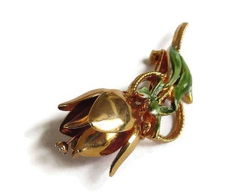 May include: A gold-toned flower brooch with green enamel leaves and a single rhinestone accent. The flower is stylized and has a rounded shape.