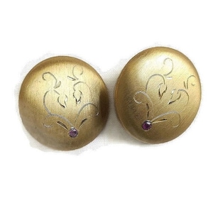 Könnte beinhalten: Ein Paar goldfarbene Ohrclips mit einem floralen Design und einem kleinen violetten Edelstein in der Mitte jedes Ohrclips.