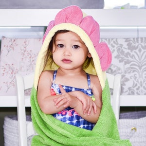 May include: A young child wearing a green and pink hooded towel with a flower design. The towel is wrapped around the child's body and the hood is pulled up over their head. The child is sitting on a white chair.