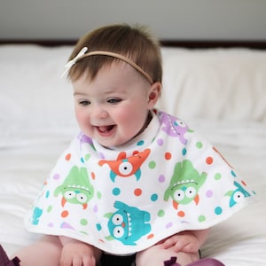 May include: A baby wearing a white bib with a colorful monster print. The bib has green, orange, purple, and blue monsters on a white background with white polka dots. The baby is wearing a white headband with a bow.