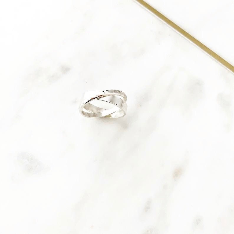 May include: A silver ring with a unique, twisted design. The ring is made of two bands that are intertwined and have a hammered texture.