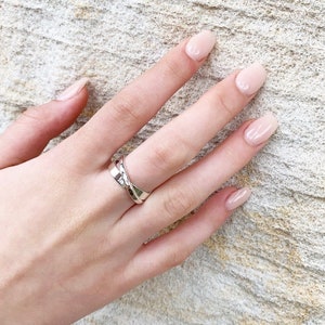 May include: A silver ring with a twisted design. The ring is worn on the middle finger of a hand with light pink nail polish.