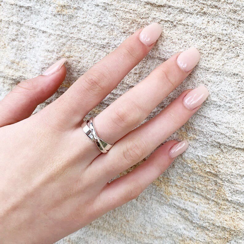 May include: A silver ring with a twisted design. The ring is worn on the middle finger of a hand with light pink nail polish.
