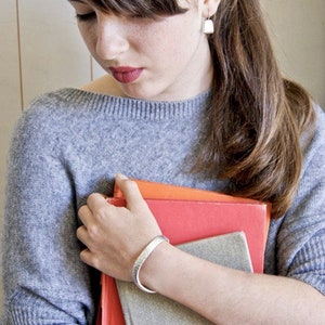 May include: A woman wearing a gray sweater holds a stack of books. She is wearing a silver bracelet on her wrist.