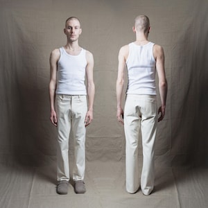 Può includere: Vista frontale e posteriore di una persona che indossa una canotta bianca a coste e pantaloni bianco sporco. I pantaloni hanno un classico design a cinque tasche. La persona indossa anche zoccoli grigi. Lo sfondo è beige neutro.