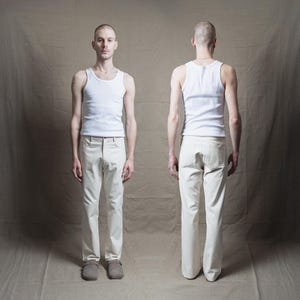 May include: A front and back view of a person wearing a white ribbed tank top and off-white pants. The pants have a classic five-pocket design. The person is also wearing gray clogs. The background is a neutral tan.