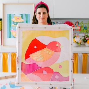 May include: A woman holds up a framed piece of fabric art featuring a pink and yellow bird with red accents on a yellow background. The bird is surrounded by pink and purple shapes.
