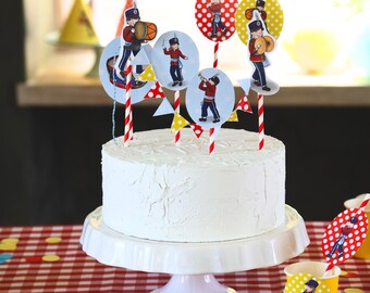 Puede incluir: Un pastel blanco decorado con banderas rojas y amarillas y adornos redondos con ilustraciones de niños tocando instrumentos musicales. El pastel está sobre un soporte de pastel blanco.