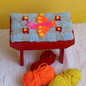 Puede incluir: Un taburete de madera rojo con un asiento acolchado azul y gris con un diseño de pájaro colorido. El taburete está rodeado de ovillos de lana en rojo, naranja y amarillo.