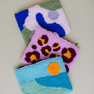 May include: Four colorful, hand-stitched pouches with different patterns. The pouches are made of a soft, fluffy material and have a zipper closure. The patterns include abstract shapes, animal print, and a sun with a blue and green landscape.