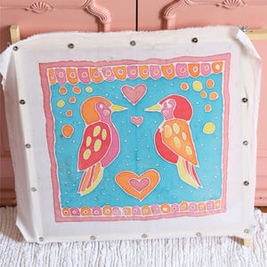 May include: A white fabric stretched on a wooden frame with two colourful birds facing each other. The birds are pink, orange, and yellow with black outlines. They are surrounded by a blue background with pink, orange, and yellow hearts and circles.