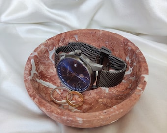 Puede incluir: Un plato de mármol rojo y blanco con un reloj de malla negra y dos anillos de oro.