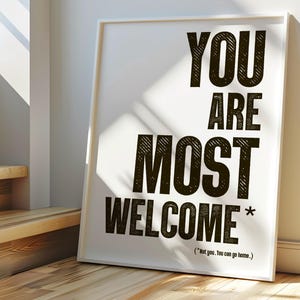 Pode incluir: Uma impressão emoldurada branca com as palavras "YOU ARE MOST WELCOME" em letras pretas. A impressão está encostada em uma parede branca em um chão de madeira, com luz solar entrando por uma janela. O texto inclui um asterisco.