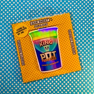 Könnte beinhalten: Ein holografischer King Pot Noodle Magnet-Lesezeichen auf einer gelben Karte mit dem Text "Sam Sweet Fabulous!" und "Home & Handmade By Me". Das Lesezeichen befindet sich in einer transparenten Plastikhülle, vor einem blauen Hintergrund mit Punkten.