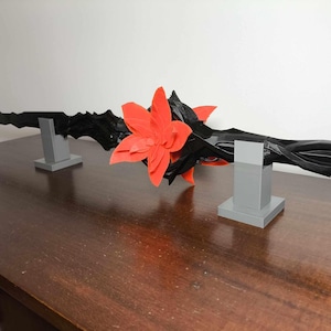 May include: A black, decorative, stylized sword-like object with a red flower in the center, resting on two gray rectangular stands. The object has a dark, textured appearance and is displayed on a wooden surface.