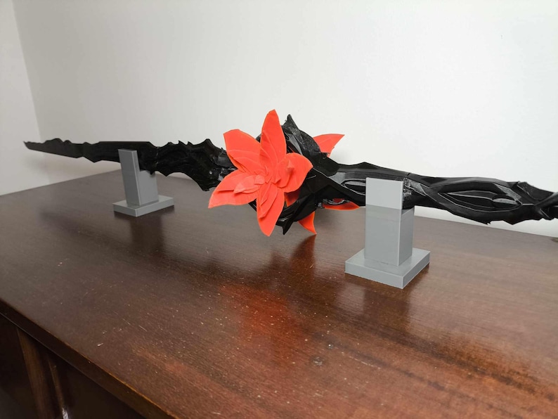 May include: A black, decorative, stylized sword-like object with a red flower in the center, resting on two gray rectangular stands. The object has a dark, textured appearance and is displayed on a wooden surface.