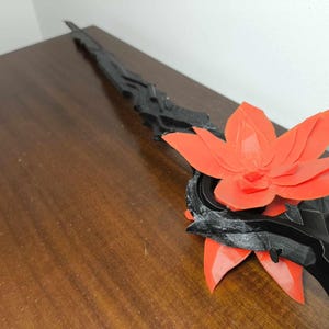 May include: A black and red decorative sword with a floral design. The sword features a black handle and blade with a large, vibrant red flower accent. The sword is displayed on a wooden surface.