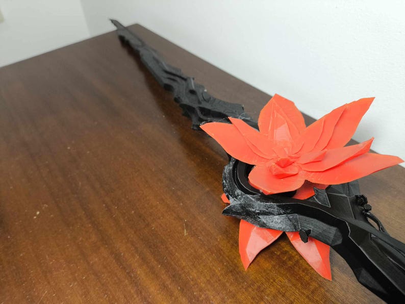 May include: A black and red decorative sword with a floral design. The sword features a black handle and blade with a large, vibrant red flower accent. The sword is displayed on a wooden surface.