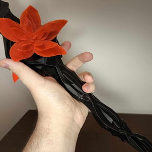May include: A black, stylized floral sculpture with a vibrant orange flower at the top. The stem has a twisted, organic design. The sculpture is held in a hand against a neutral background.