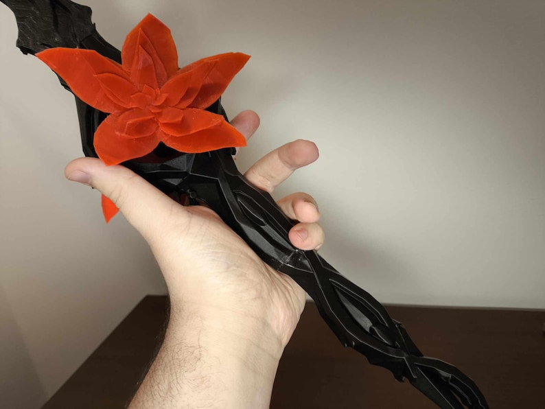 May include: A black, stylized floral sculpture with a vibrant orange flower at the top. The stem has a twisted, organic design. The sculpture is held in a hand against a neutral background.