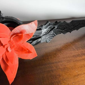 May include: A 3D-printed, black and red decorative object. A large, red flower is on the left, with a detailed, multi-layered petal design. To the right, a black, textured, spear-like element extends, suggesting a fantasy or decorative theme.