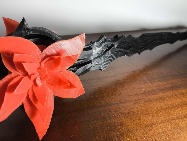 May include: A 3D-printed, black and red decorative object. A large, red flower is on the left, with a detailed, multi-layered petal design. To the right, a black, textured, spear-like element extends, suggesting a fantasy or decorative theme.