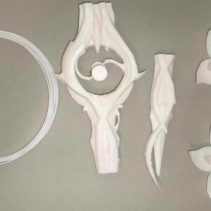May include: An assortment of white, 3D-printed decorative elements. The set includes a circular ring, a stylized central piece with an eye-like design, a slender, curved component, and two flower-shaped pieces. The items are arranged on a gray surface.
