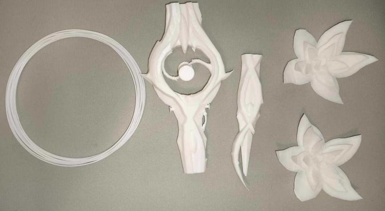 May include: An assortment of white, 3D-printed decorative elements. The set includes a circular ring, a stylized central piece with an eye-like design, a slender, curved component, and two flower-shaped pieces. The items are arranged on a gray surface.