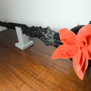May include: A black, textured, blade-like object is displayed on a gray stand, with a large, orange flower on the right. The objects are on a brown wooden surface. The blade has a jagged edge.